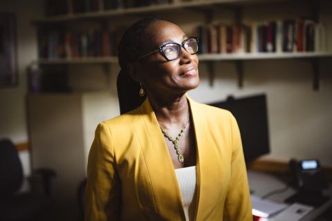 Simone A James Alexander poses for a photo in a yellow blazer. She looks towards the light and smiles softly. Behind her are book shelves.